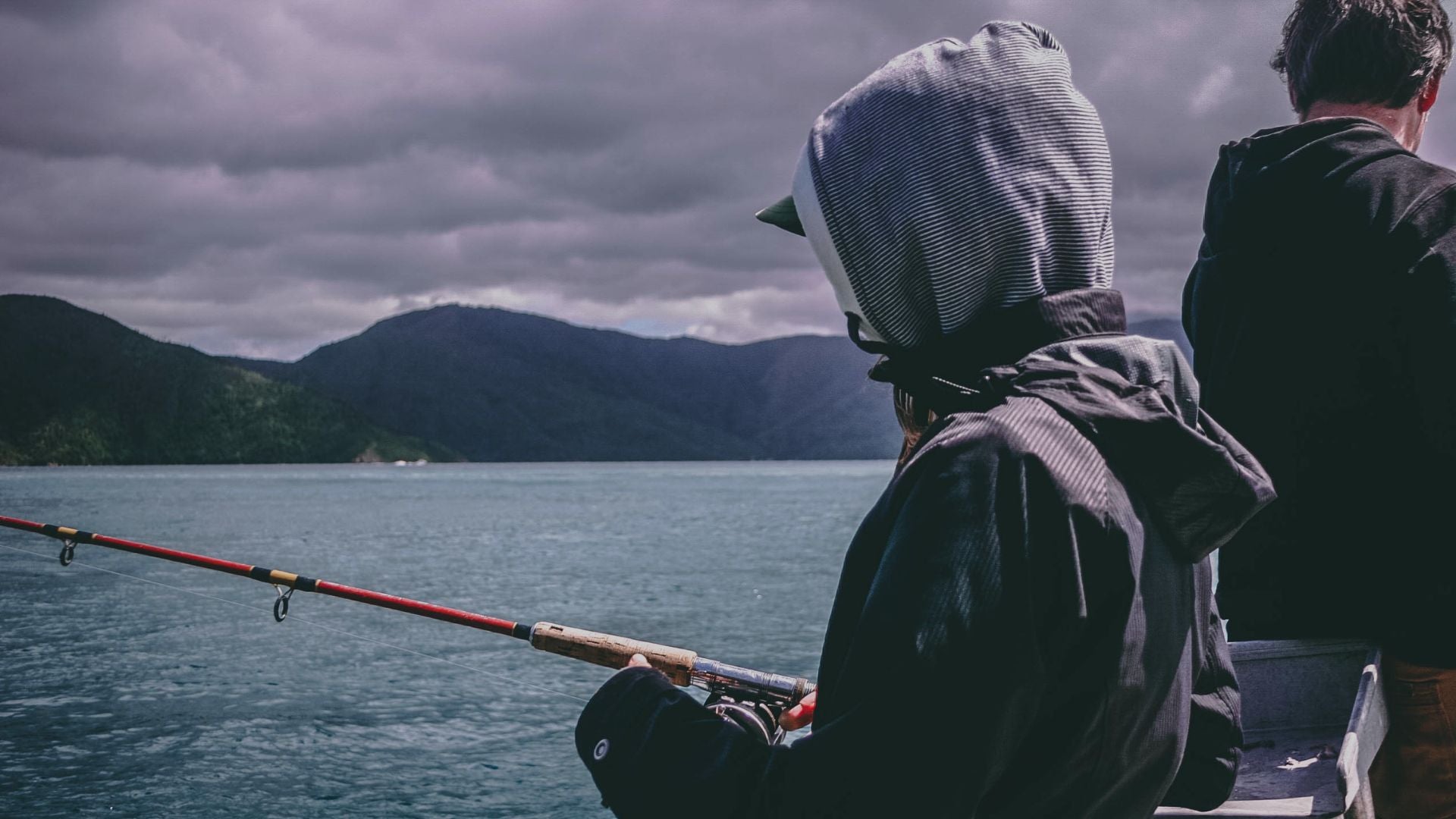 Two people fishing on a boat with mountains and cloudy sky in the background