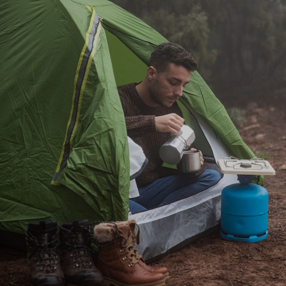 Man camping, sitting inside a green tent and preparing a drink.