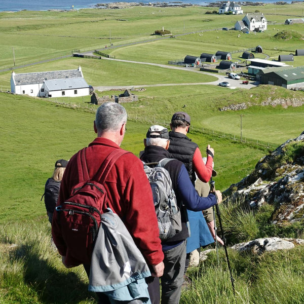 Group of people hiking with backpacks on a grassy landscape with buildings in the distance.