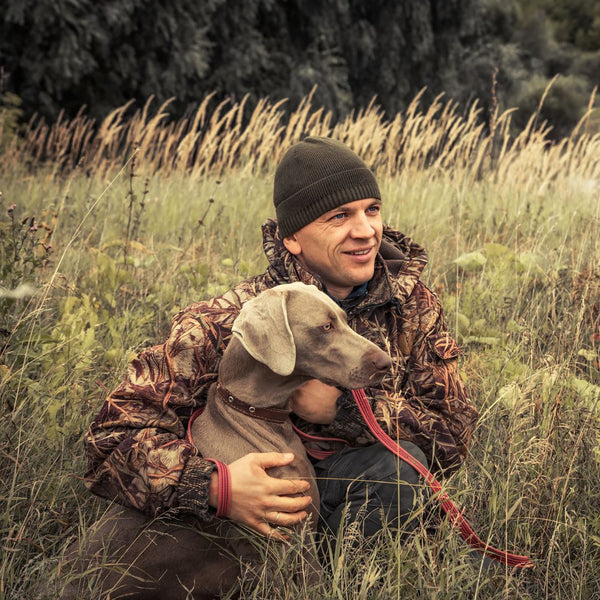 Man in camouflage jacket and beanie sitting in a field with a dog on a leash