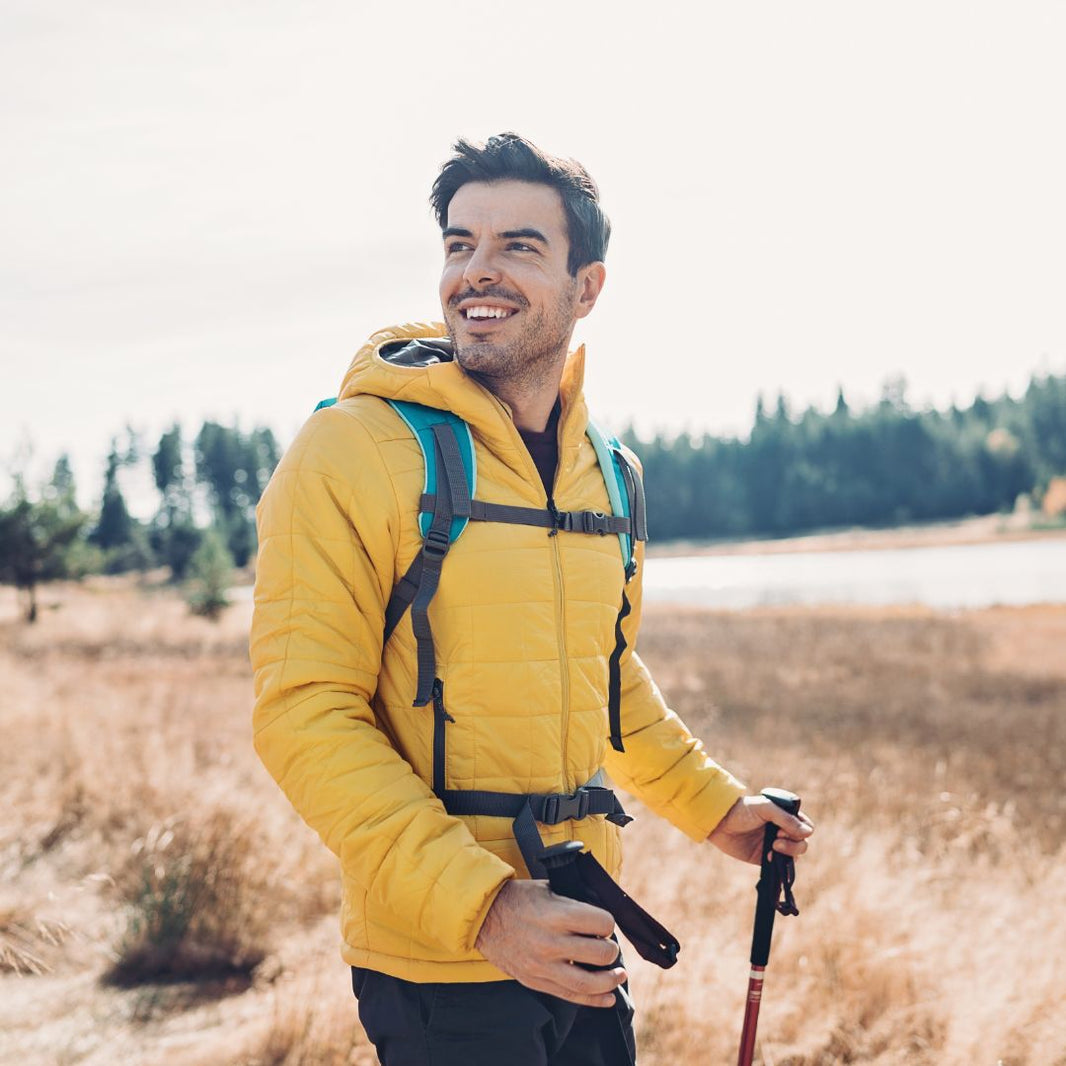 Man in yellow jacket with backpack and hiking poles standing in a field with trees in the background